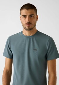 Minzgrünes T-Shirt mit kurzen Ärmeln aus strukturiertem Material. Verfügt über einen Rundhalsausschnitt und ein kleines schwarzes Logo auf der linken Brust.