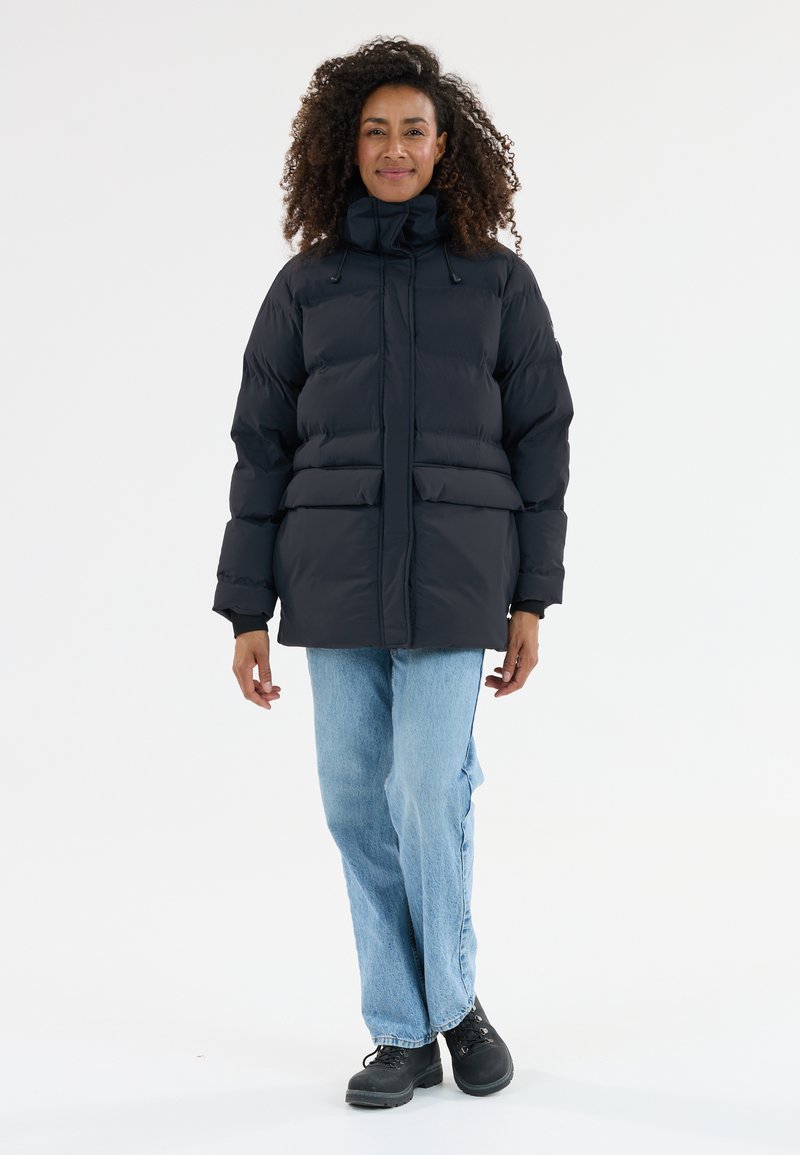 Dunkle Pufferjacke mit hohem Kragen, frontalen Taschen und lockerer Passform. Kombiniert mit hellblauen Jeans und schwarzen Schnürstiefeln.