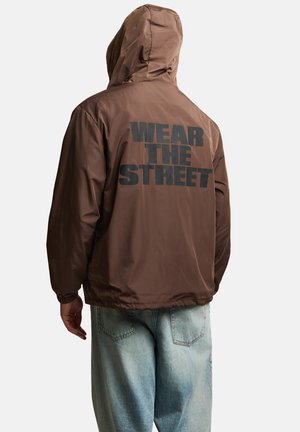 Osoba nosząca brązową kurtkę z kapturem z napisem "WEAR THE STREET" dużymi czarnymi literami na plecach, w połączeniu z jasnoniebieskimi jeansami.