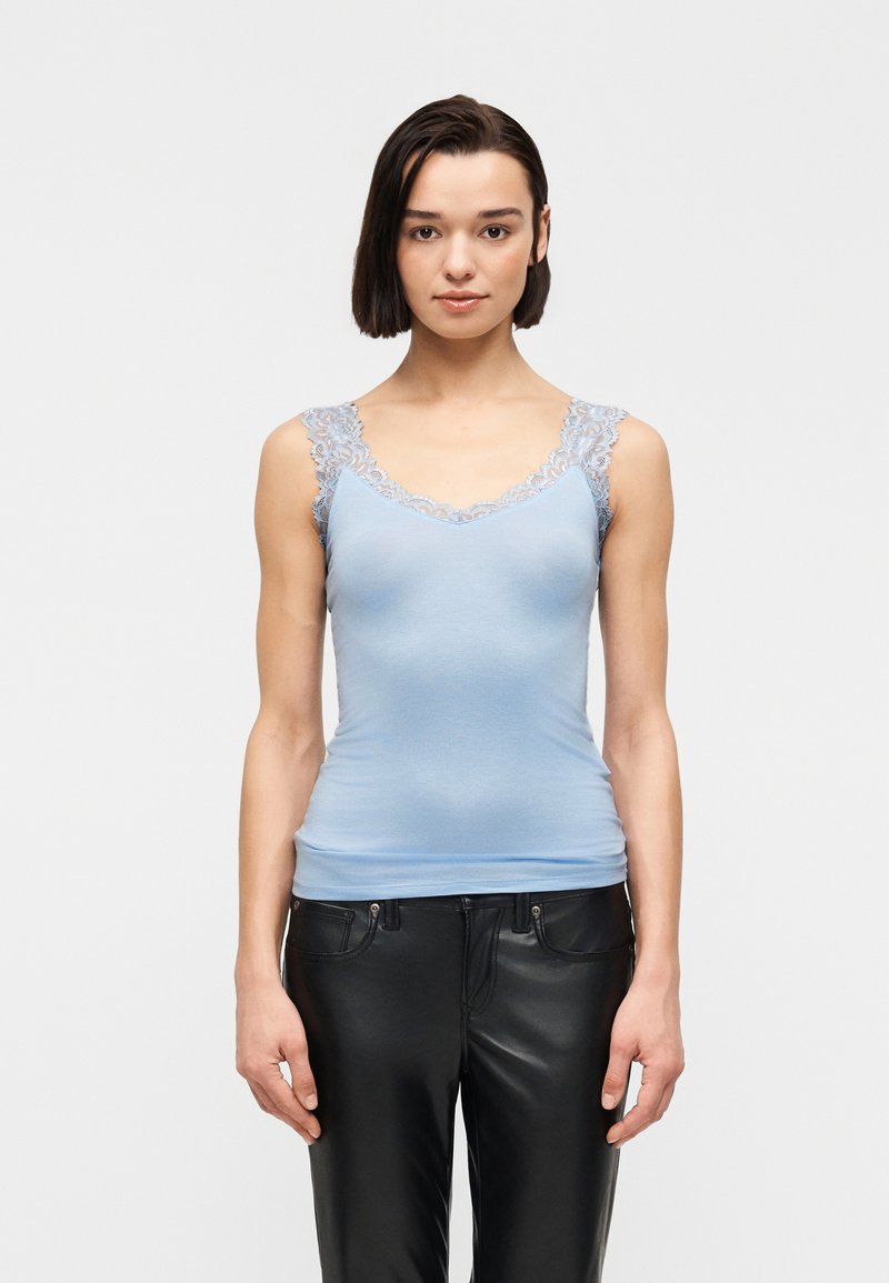 Γυναίκα με κοντά σκούρα μαλλιά, ντυμένη με ανοιχτό μπλε τοπ με δαντέλα και μαύρο δερμάτινο παντελόνι, που στέκεται μπροστά σε λευκό, απλό φόντο.