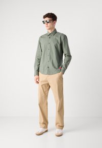 Cămașă verde închis cu guler și închidere cu nasturi, asortată cu pantaloni bej și adidași albi. Ochelarii de soare completează ținuta.