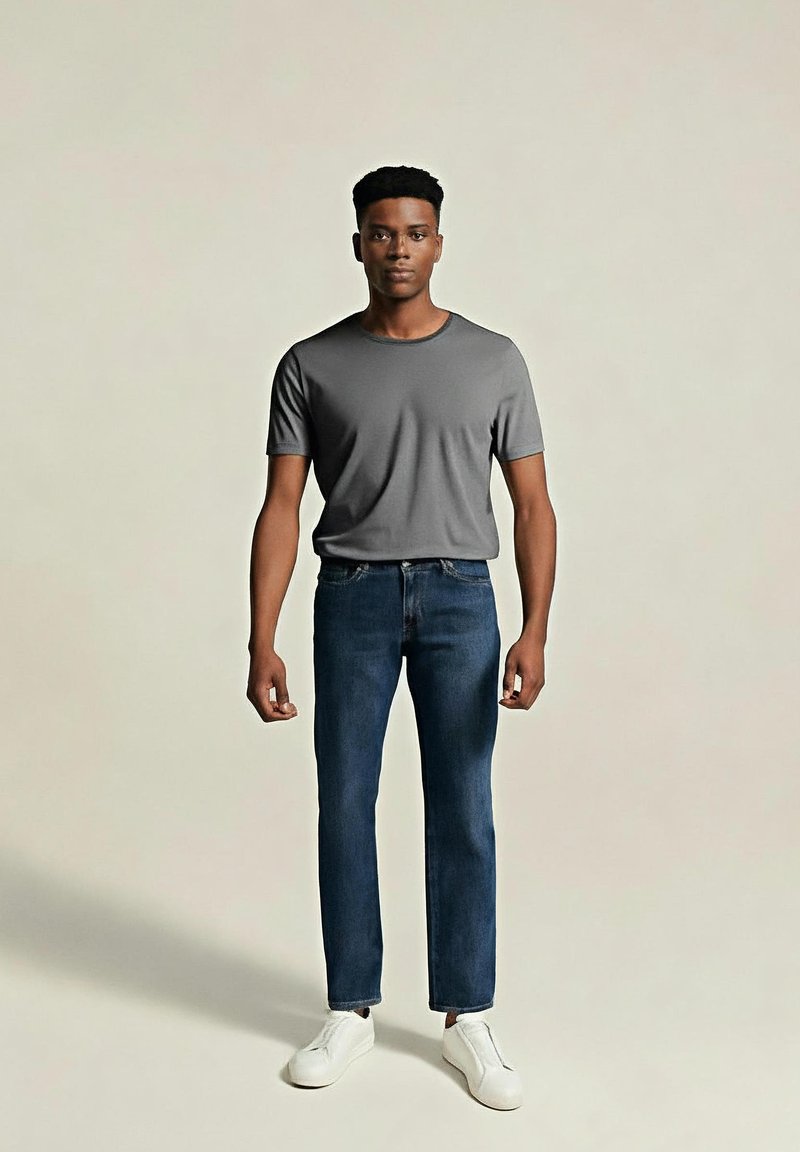 Junger Mann steht mit dem Gesicht nach vorne, trägt ein graues T-Shirt, dunkeblaue Jeans und weiße Slipper gegen einen einfarbigen Hintergrund.