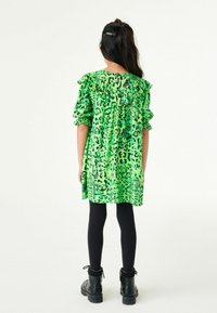 Fille aux longs cheveux bruns portant une robe verte à imprimé léopard, des leggings noirs et des bottes noires, se tenant de dos sur un fond blanc.