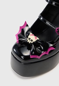 Căciulă din piele patent neagră, cu platformă groasă, având un fundiță în formă de lilieci roz și negru și un accent cu un craniu și fundițe roz.