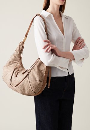 Borsa hobo beige con texture, dalla forma morbida, dettagli in pelle marrone, chiusure con zip e tracolla. Portata da una persona che indossa una blusa bianca.