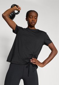 T-shirt de sport noir à manches courtes avec un détail froncé sur le côté, associé à un legging noir. Le matériau semble léger et lisse.