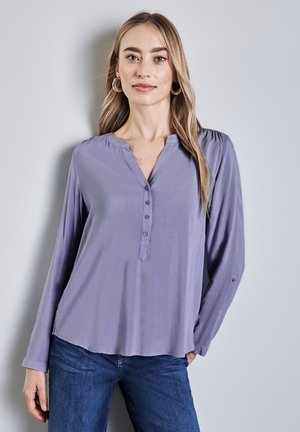 Blusa a maniche lunghe lilla con scollatura a V e patta con bottoni, caratterizzata da pieghe sulle spalle e una vestibilità comoda, abbinata a jeans blu.