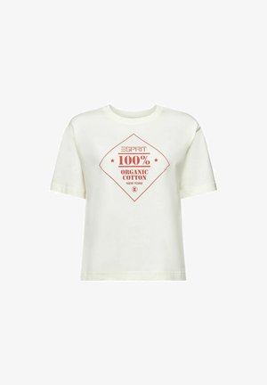 Person iført en hvid T-shirt med rød tekst "ESPRIT 100% Organic Cotton New York", blå jeans og en stribet sweater bundet omkring taljen.