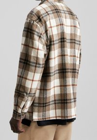 Osoba w brązowej, białej i czarnej koszuli w kratę z długim rękawem, założonej na czarną koszulkę, oraz w beżowych spodniach, stoi tyłem do aparatu.