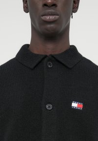 Μαύρο πλεκτό ζακέτα με κουμπωτή πατιλέτα και γιακά. Διαθέτει κονκάρδα με το λογότυπο Tommy Jeans σε μπλε, κόκκινο και λευκό. Μαλακή υφή.