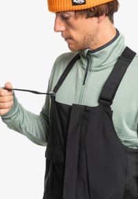 Unos tirantes negros con peto ajustables, llevados sobre una camiseta texturizada de color verde claro. Los detalles notables incluyen bolsillos frontales y herrajes de sujeción.