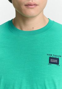 Zöld póló puha anyagból, környakú, rövidujjú, elöl bal oldalon kis címkével, amin az áll: "TOM TAILOR".