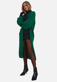 Cardigan lungo verde in maglia con maniche oversize, caratterizzato da un motivo texturizzato e due tasche frontali, indossato sopra un vestito nero e collant.