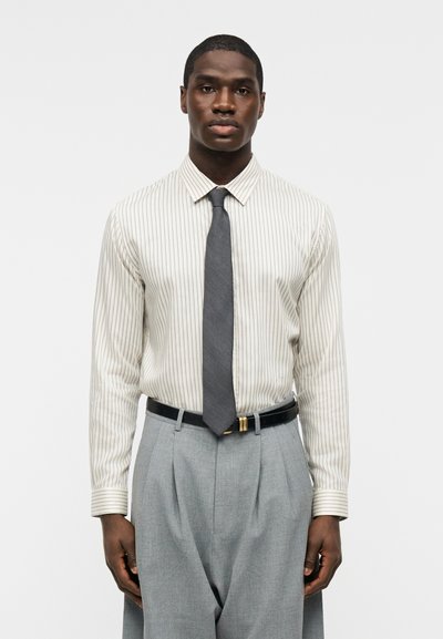 Άνδρας με άσπρο πουκάμισο με λεπτές κάθετες ρίγες, γκρι γραβάτα, μαύρη ζώνη και ανοιχτό γκρι πλισέ παντελόνι, στέκεται μπροστά σε απλό φόντο.