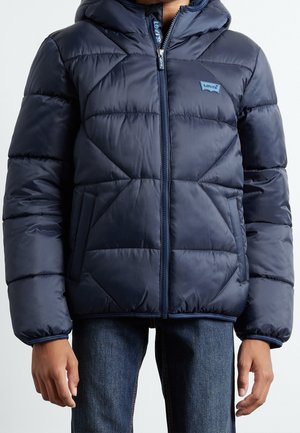Enfant portant une veste matelassée bleu marine avec une fermeture éclair à l'avant, montrant le logo Levi's sur la poitrine, et un jean bleu.