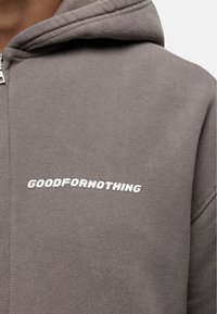 Grijze zip-up hoodie van zacht materiaal, met opvallende witte tekst "GOODFOR NOTHING" op de linkerkant van de borst. Gladde textuur.