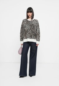 Žena nosi duksericu s uzorkom leopard, široke tamne traperice, crne cipele i drži malu svijetlopurple torbicu na bijeloj pozadini.