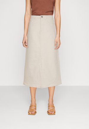 Woman wearing a beige midi skirt, rust-colored sleeveless top, and tan strappy square-toe heels standing against a plain background.