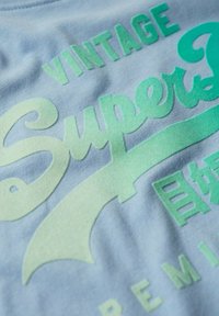  Ljusblå bomulls-T-shirt med gradient gröna texter: "Vintage SuperDry" med stiliserad typografi och subtila texturer.