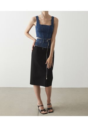 Kobieta w bezrękawnym, dżinsowym topie odsłaniającym brzuch i czarnej, midi spódnicy z przednim zamkiem błyskawicznym, zestawionej z czarnymi sandałami na obcasie na jednolitym tle.