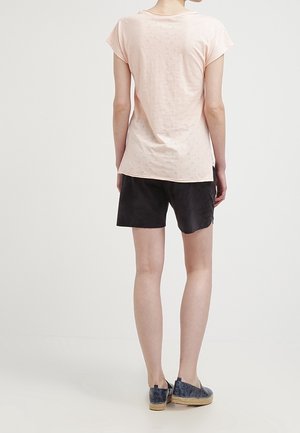 Personne portant un haut rose clair à manches courtes, un short noir et des chaussures foncées sans lacets, debout, tournée de dos sur un fond uni.