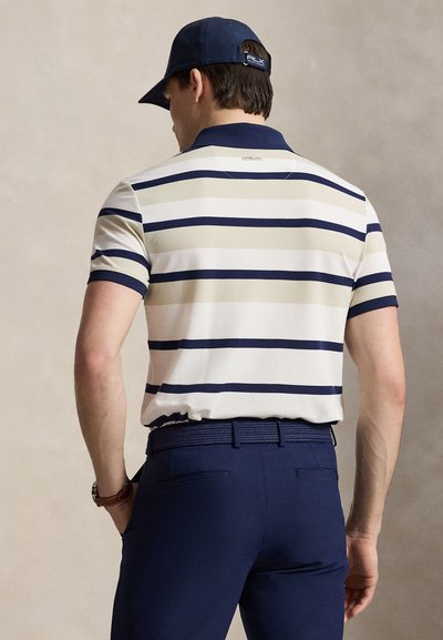 Homme portant une casquette bleu marine, un pantalon bleu marine avec ceinture, et un polo à manches courtes avec des rayures horizontales bleu marine et beige, vu de dos.