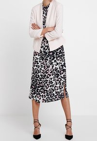 Jasnoróżowa marynarka na czarno-białej sukience w panterkę z różowymi akcentami, z bocznymi rozcięciami. Całość dopełniają czarne sandały na obcasie z paskami.