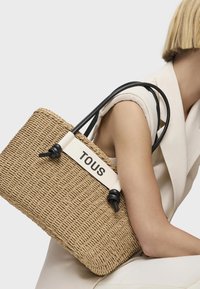 Bolso de tote tejido de paja natural con asas de cuero negro y una banda blanca que muestra el nombre de la marca "TOUS". Superficie texturizada, forma rectangular.