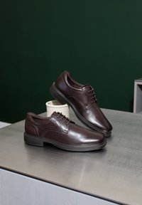 Paar polierte dunkelbraune Leder-Schnürschuhe, dargestellt auf einer Metalloberfläche mit dunkelgrünem Hintergrund.