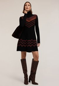 Abito a maglia nero con motivi a zigzag in arancione ruggine, colletto a dolcevita, gonna svasata, abbinato a stivali alti fino al ginocchio in pelle di serpente marrone e una borsa in pelliccia.