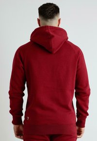 Sudadera roja hecha de tela suave. Cuenta con una capucha ajustable con cordón y puños de canalé. Diseño sencillo con un pequeño detalle de logo cerca del dobladillo.