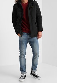 Svart huva jacka med faux pälsdetalj, frampocketar och dragsko, bärs över en burgundy sweatshirt och ljusblå jeans. Svarta sneakers.
