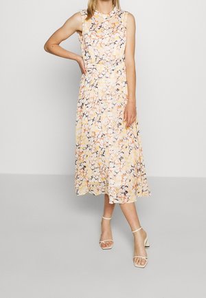 Femme portant une robe fleurie sans manches, longue jusqu'à la cheville, avec des motifs jaune pastel, orange et noir, et des sandales blanches à talons avec brides à la cheville.