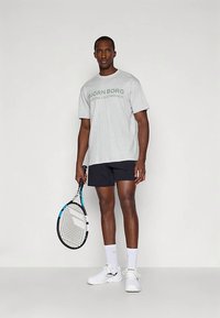 Pelēka T-krekls ar zaļu tekstu, melnas šorti, balti apavi un balti zeķes. Tur rokā tenisa raketi ar zilmelnu rakstu.