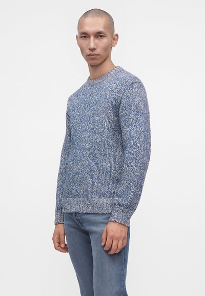 Homme portant un pull en laine bleu et gris et un jean bleu, debout devant un fond blanc uni.