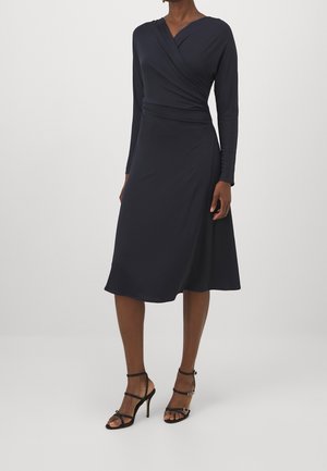 Femme portant une robe portefeuille bleu marine à manches longues et longueur genou, accompagnée de sandales noires à talons hauts, sur un fond clair uni.