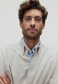 En person med lockigt hår och skägg bär en ljusbeige skjorta med knappar över en blårandig skjorta med krage, med armarna korsade.