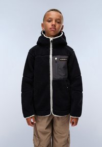 Veste en polaire noire avec capuche et bord blanc, dotée d'une poche zippée à l'avant en gris. Portée sur un pantalon beige, avec un arrière-plan clair.