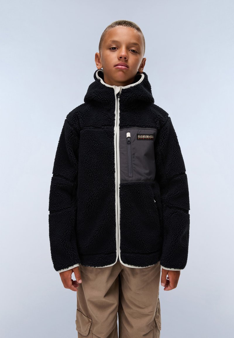 Veste en polaire noire avec capuche et bord blanc, dotée d'une poche zippée à l'avant en gris. Portée sur un pantalon beige, avec un arrière-plan clair.