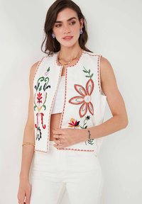 Witte mouwloze vest met kleurrijke bloemenborduursels, omrand met rode stiksel en een ronde halsontwerp.