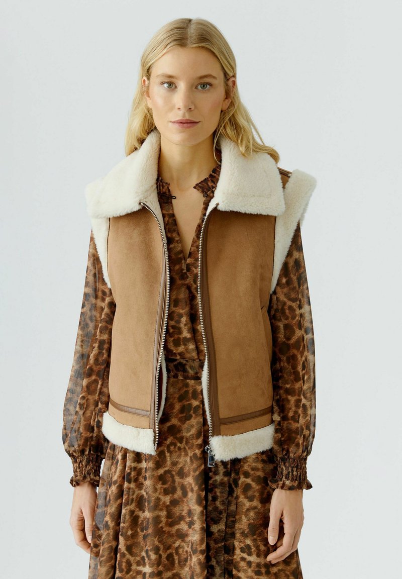 Blonde Frau, die eine braune Kunstfellweste über einem langärmeligen Leopardenmusterkleid vor einem einfarbigen hellen Hintergrund trägt.