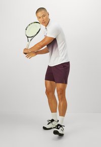Homme en tenue de tennis blanche et violette tenant une raquette, prêt à frapper un coup de revers sur un fond uni.