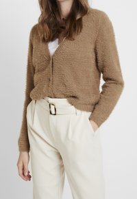 Brun fluffig cardigan i mjukt material, med knäppning och avslappnad passform, i kombination med krämfärgade högmidjade byxor.