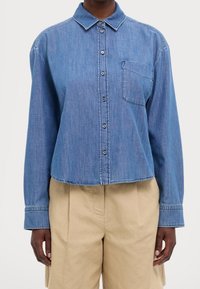 Ljusblå denimskjorta med knappar, krage, långa ärmar, en bröstficka och en kortad nederkant. Slät textur och avslappnad passform.