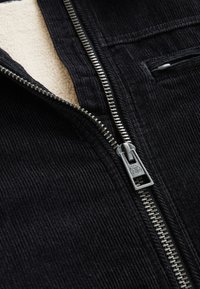 Gros plan d'une veste en velours côtelé noire partiellement dézippée avec une fermeture éclair argentée et une doublure polaire beige visible.