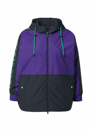 Veste coupe-vent violette et noire avec capuche, ornée d'accents verts sur les cordons de serrage et de texte le long des manches, avec une fermeture éclair.