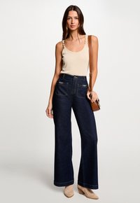 High-waisted dark blue denim flared pants with front pockets and contrast stitching, paired with a beige tank top and brown shoulder bag.