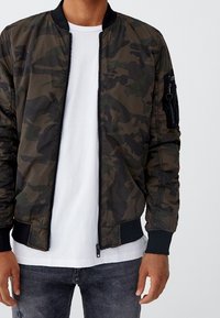Veste bomber camouflage en vert foncé et noir, dotée d'une fermeture éclair frontale, de poignets côtelés et d'une poche zippée sur la manche.