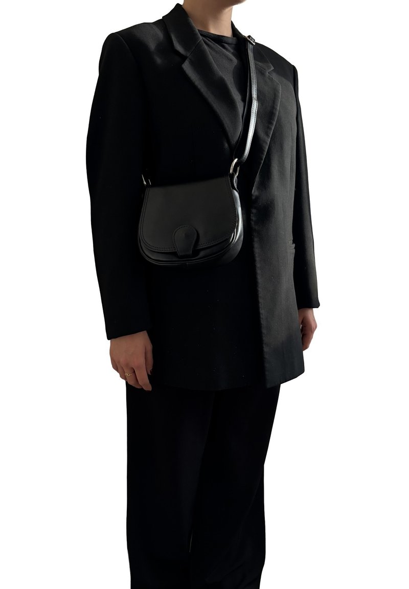Borsa a spalla in pelle nera con forma curvata, chiusura a pattina e tracolla regolabile, indossata sopra un blazer nero e pantaloni abbinati.