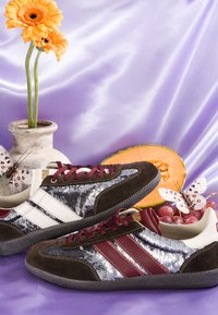 Deux baskets à paillettes avec des rayures bordeaux et blanches sur une surface en soie violette, entourées de fleurs jaunes, d'une tranche de melon, de raisins et de papillons décoratifs.